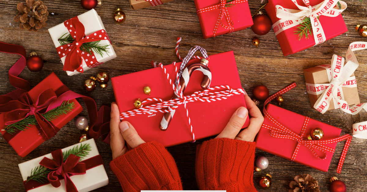 Hands holding a wrapped red holiday gift surrounded by festive boxes, supporting the Hash House A Go Go article on seasonal gift card deals, comfort food gift ideas, and holiday dining promotions.